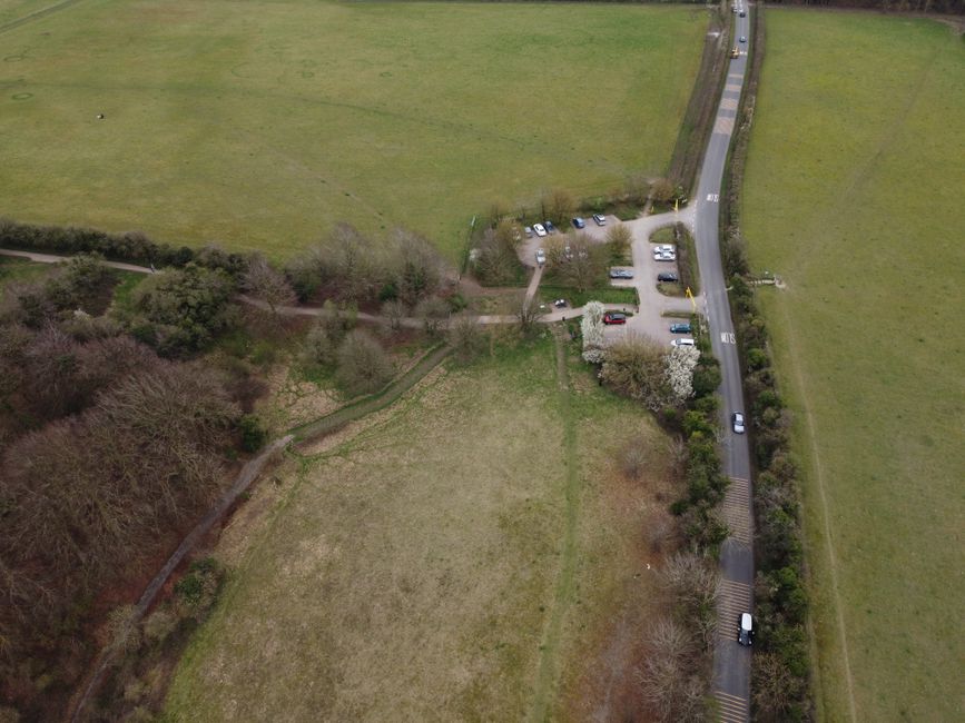 An aerial view of a green landscape showing a large grassy area intersected by a narrow dirt pathway and bordered by trees and shrubs. Adjacent to the open field is a small parking lot with several pa