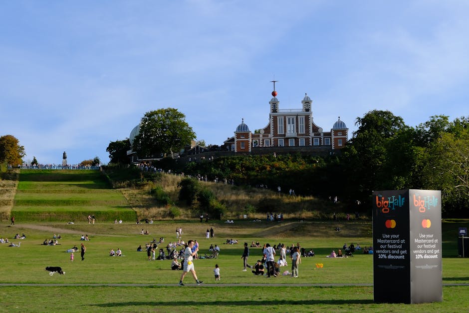 A landscaped park area with a gently sloping grassy lawn populated by various groups of people sitting, walking, and picnicking on the lush, green, well-maintained grass. The lawn features a series of