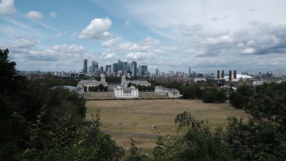 A landscaped park area with a gently sloping grassy lawn populated by various groups of people sitting, walking, and picnicking on the lush, green, well-maintained grass. The lawn features a series of